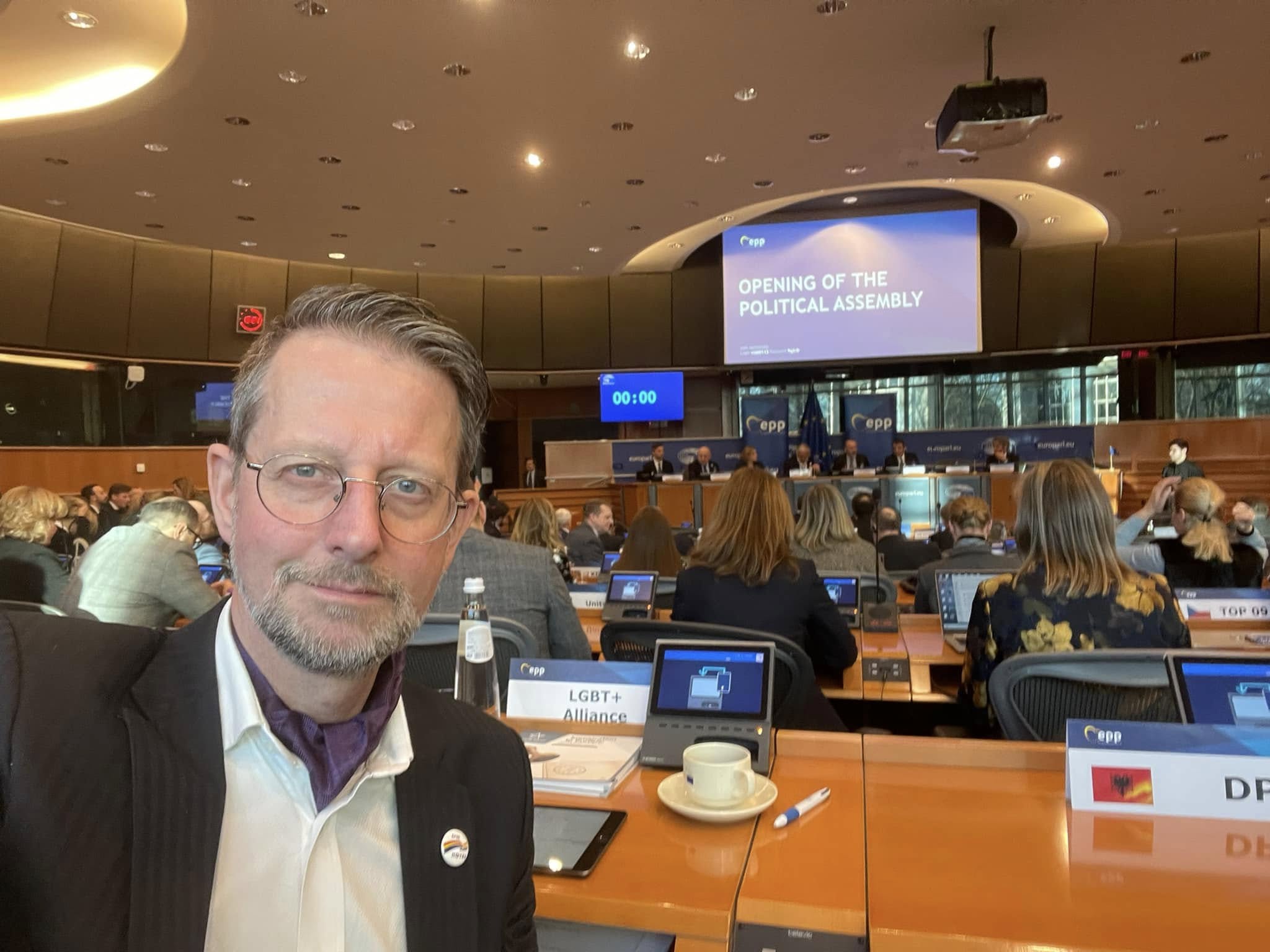 Mann i et møterom, skjerm med teksten "opening of the political assembly" i bakgrunnen.
