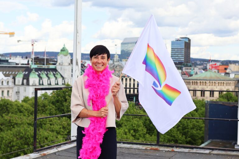 Bilde av Høyres partileder, Ine Eriksen Søreide, som holder i et supporterflagg med Høyres logo i regnbuefarger på taket av Høyres Hus i Oslo, med rosa fjørboa rundt halsen.